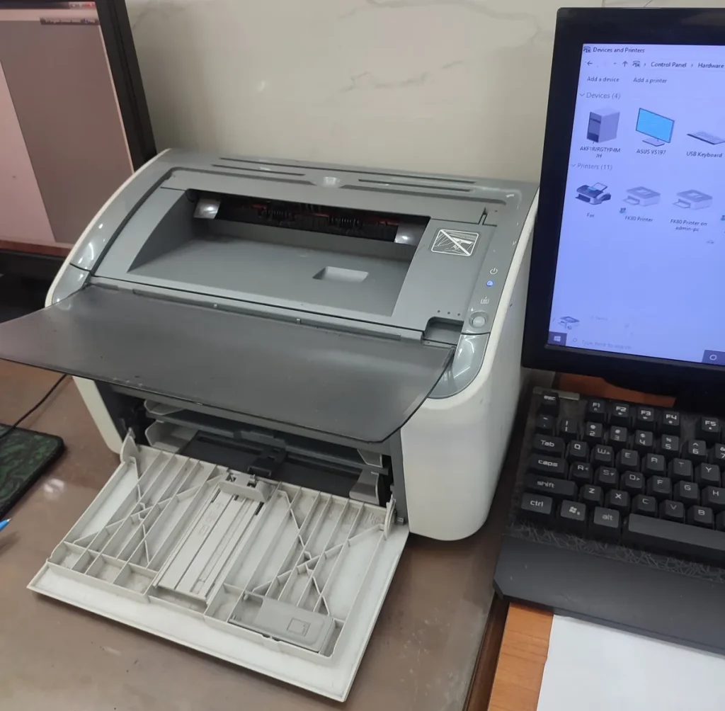 Máy in HP LaserJet mở khay giấy, bên cạnh màn hình mở cửa sổ Devices and Printers